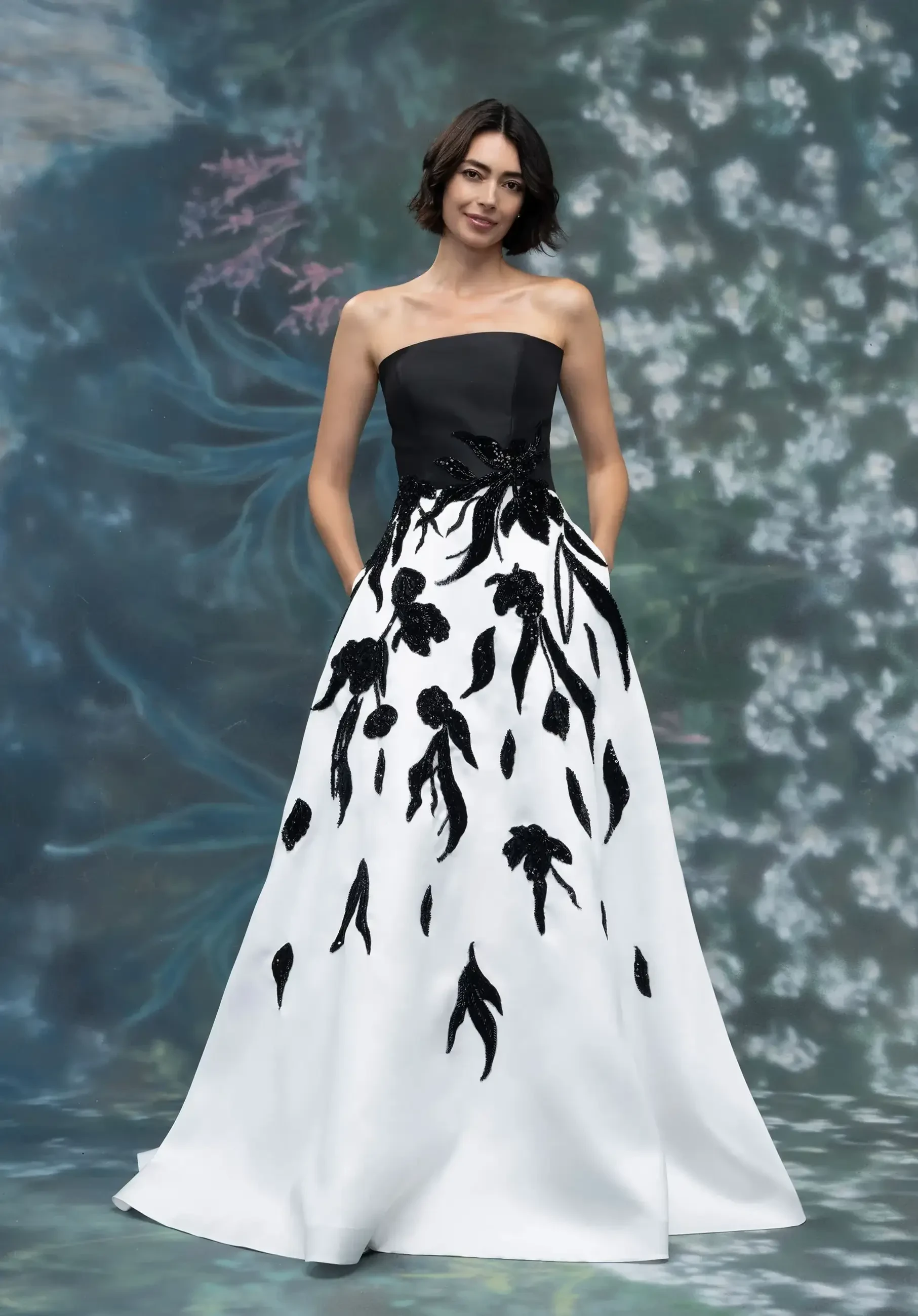 A model wearing a strapless black and white gown with floral embroidery, standing against a floral background.