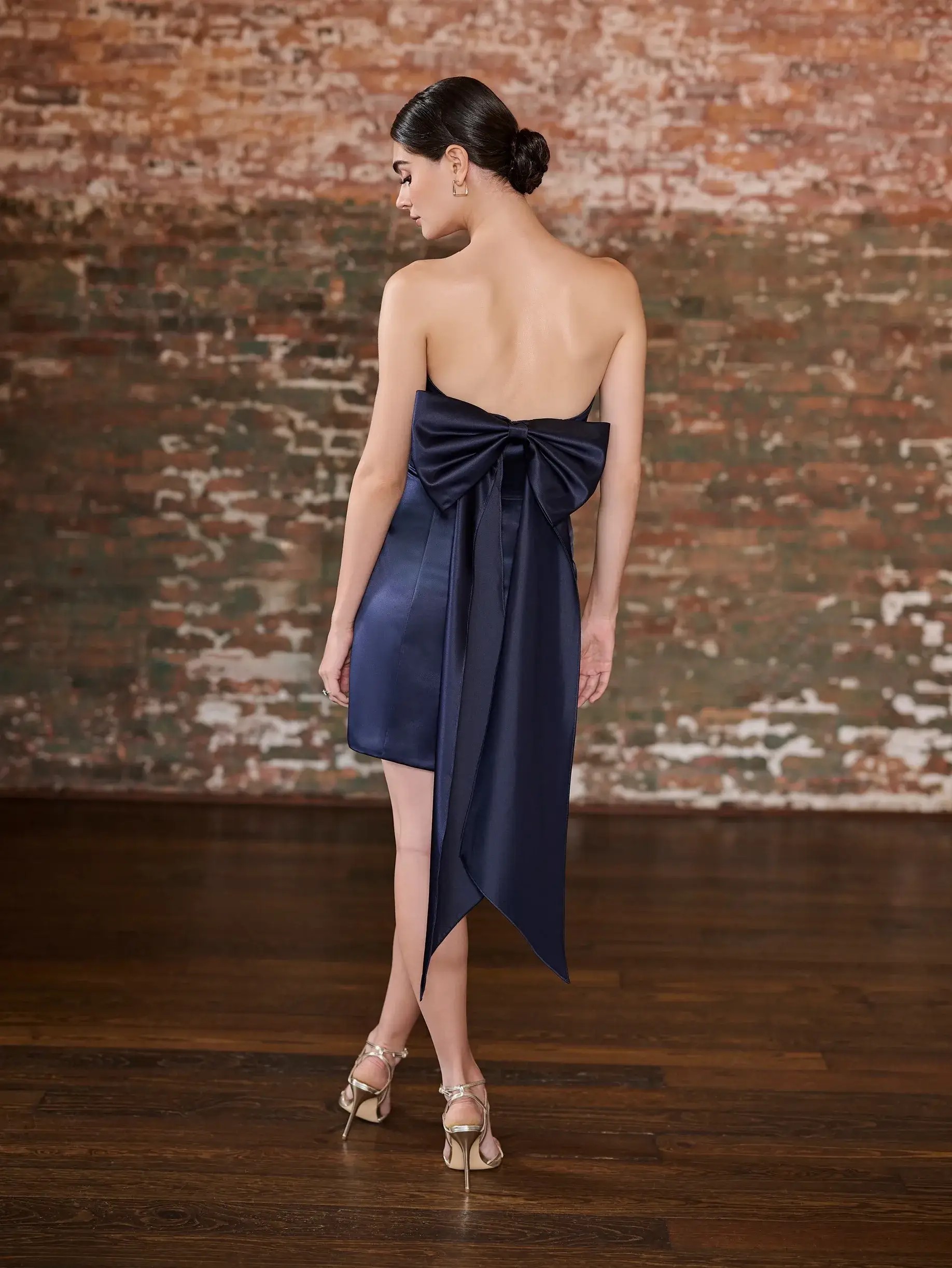 Model is wearing a short satin bridesmaid dress with an oversized bow and notched neckline