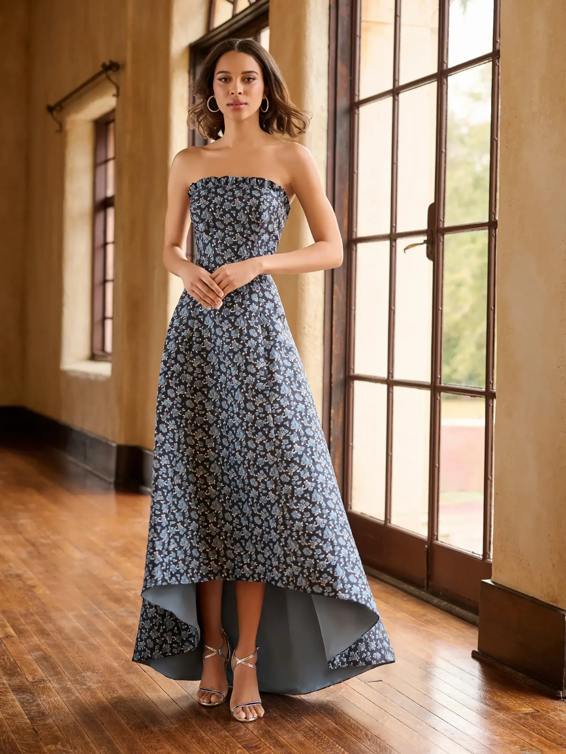 Woman in a strapless, floral high-low gown stands indoors on wooden floor, near tall windows. She exudes elegance and calm with a gentle smile.