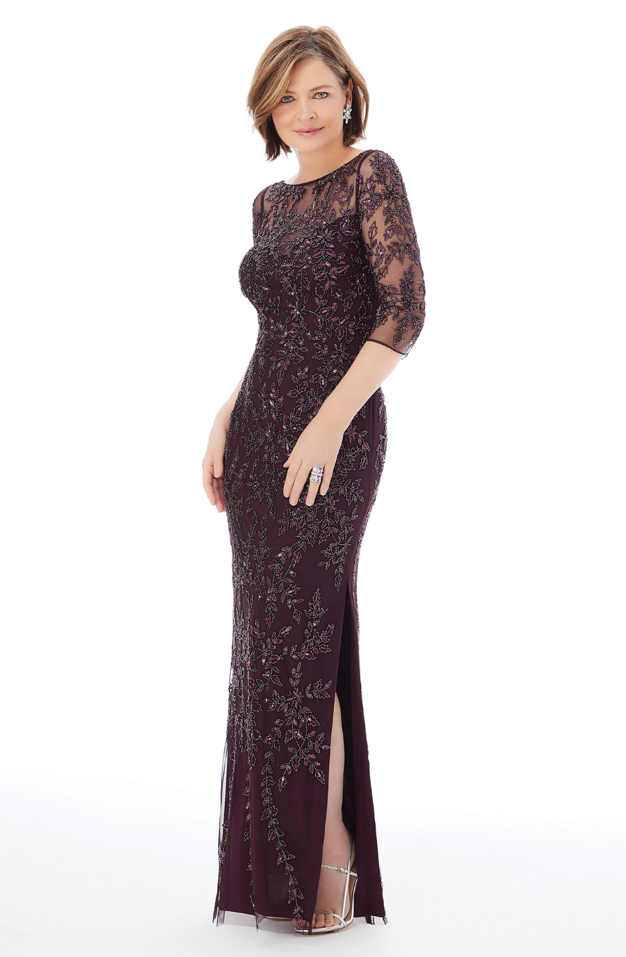A woman wearing a long, elegant burgundy dress with intricate lace detailing and three-quarter sleeves. The dress features a thigh-high slit and she is standing posed in front of a white background.