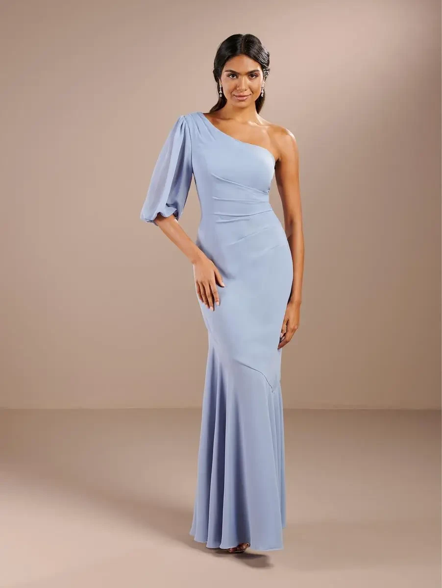 A woman in a light blue one-shoulder gown stands against a neutral background. The dress features a flowing design and captures an elegant, poised demeanor.