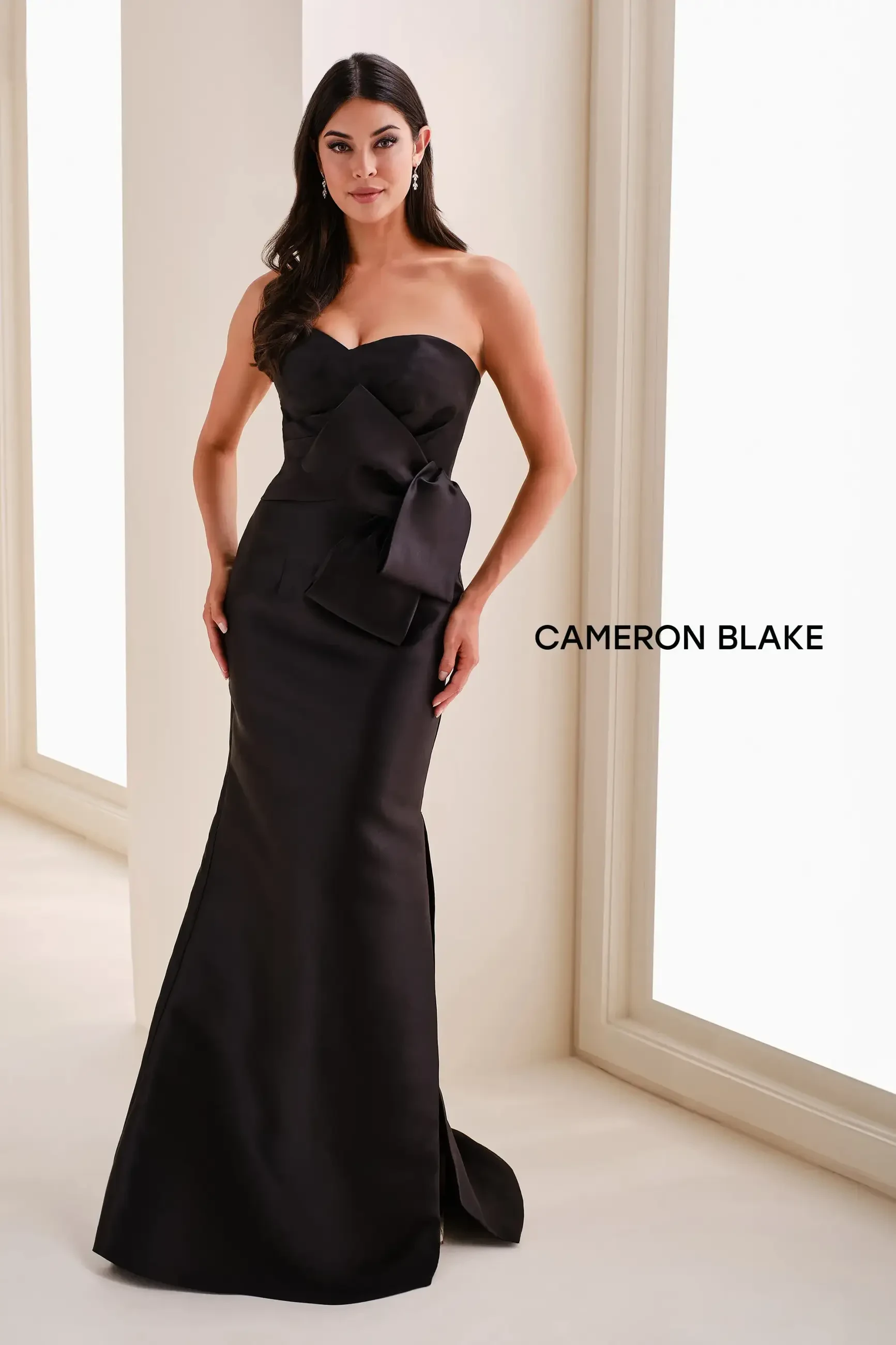 A woman in a black strapless gown with a bow detail standing against a light background. The text 