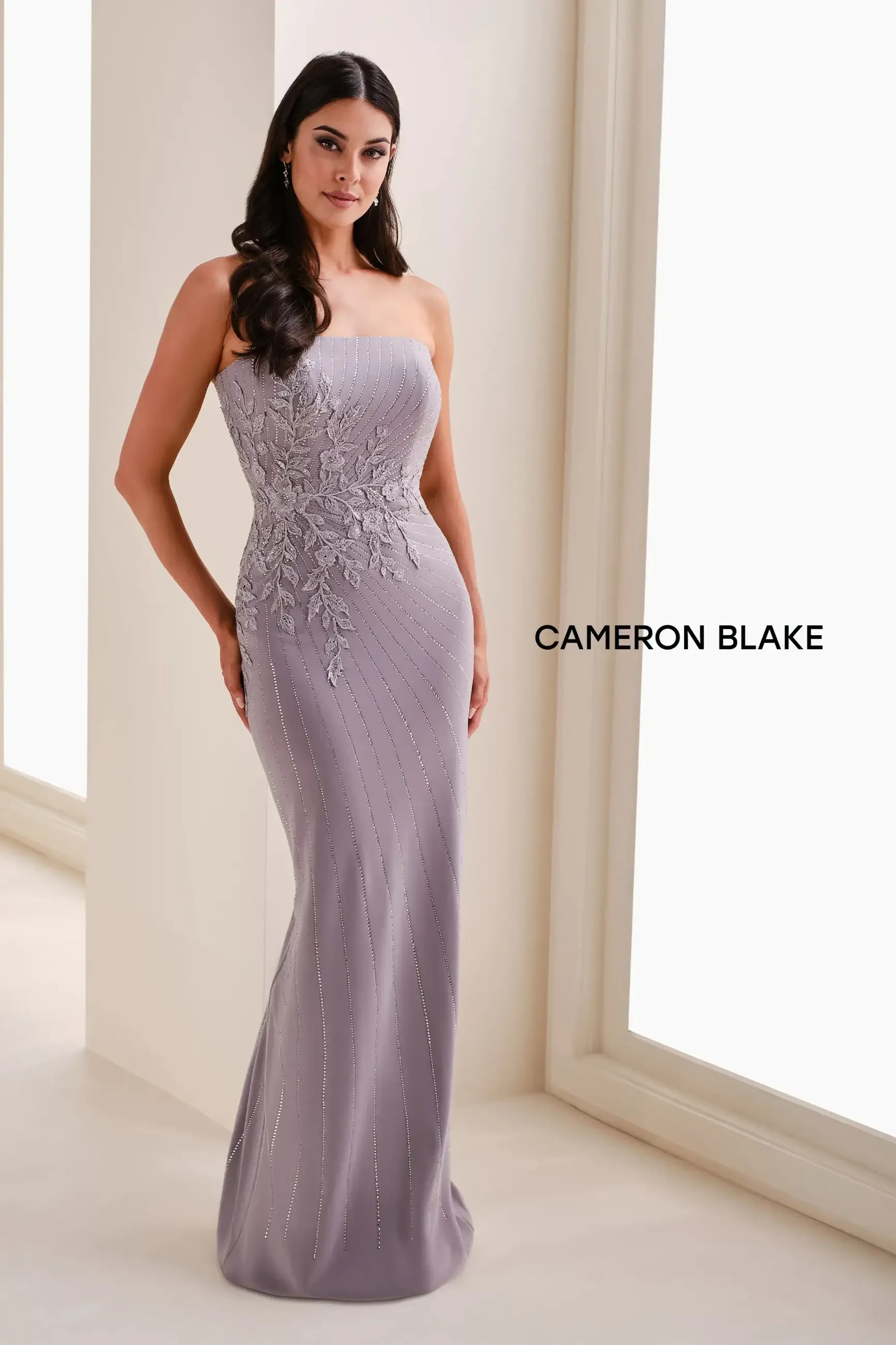 A woman wearing a strapless lavender evening gown with intricate embroidery on one side, standing in a well-lit space. The dress is fitted and flows down to the floor.