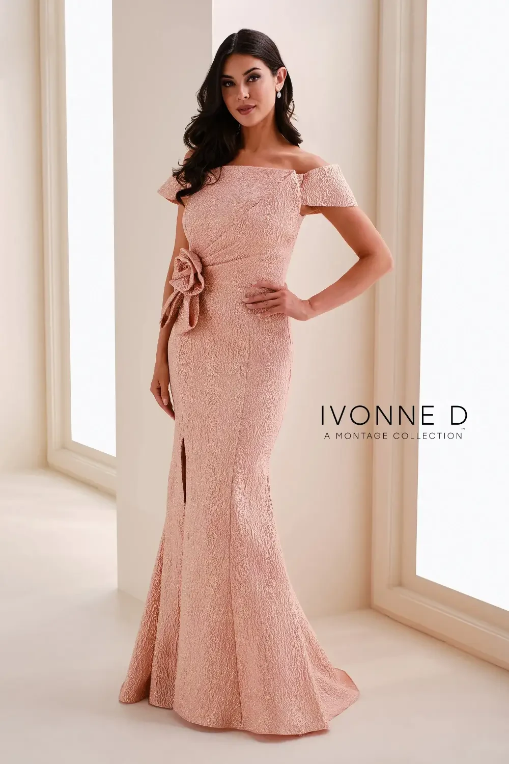 A woman wearing a pink, off-the-shoulder evening gown with a fitted silhouette and a slit, posing in a well-lit room.