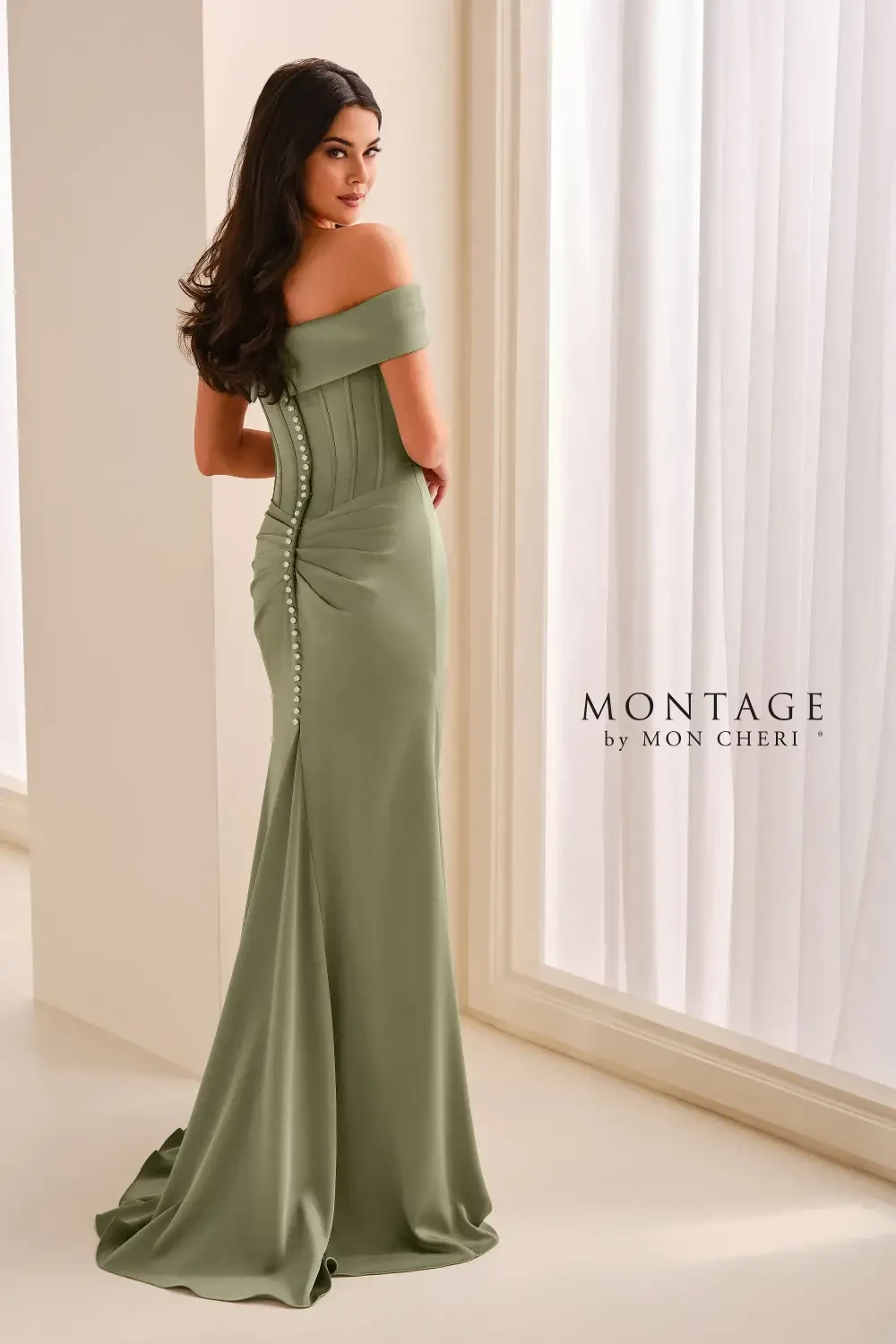 A woman wearing a form-fitting, off-the-shoulder, olive-green evening gown with a sleek design and button detailing along the back. She is positioned with her back to the camera, standing in a well-lit interior space.