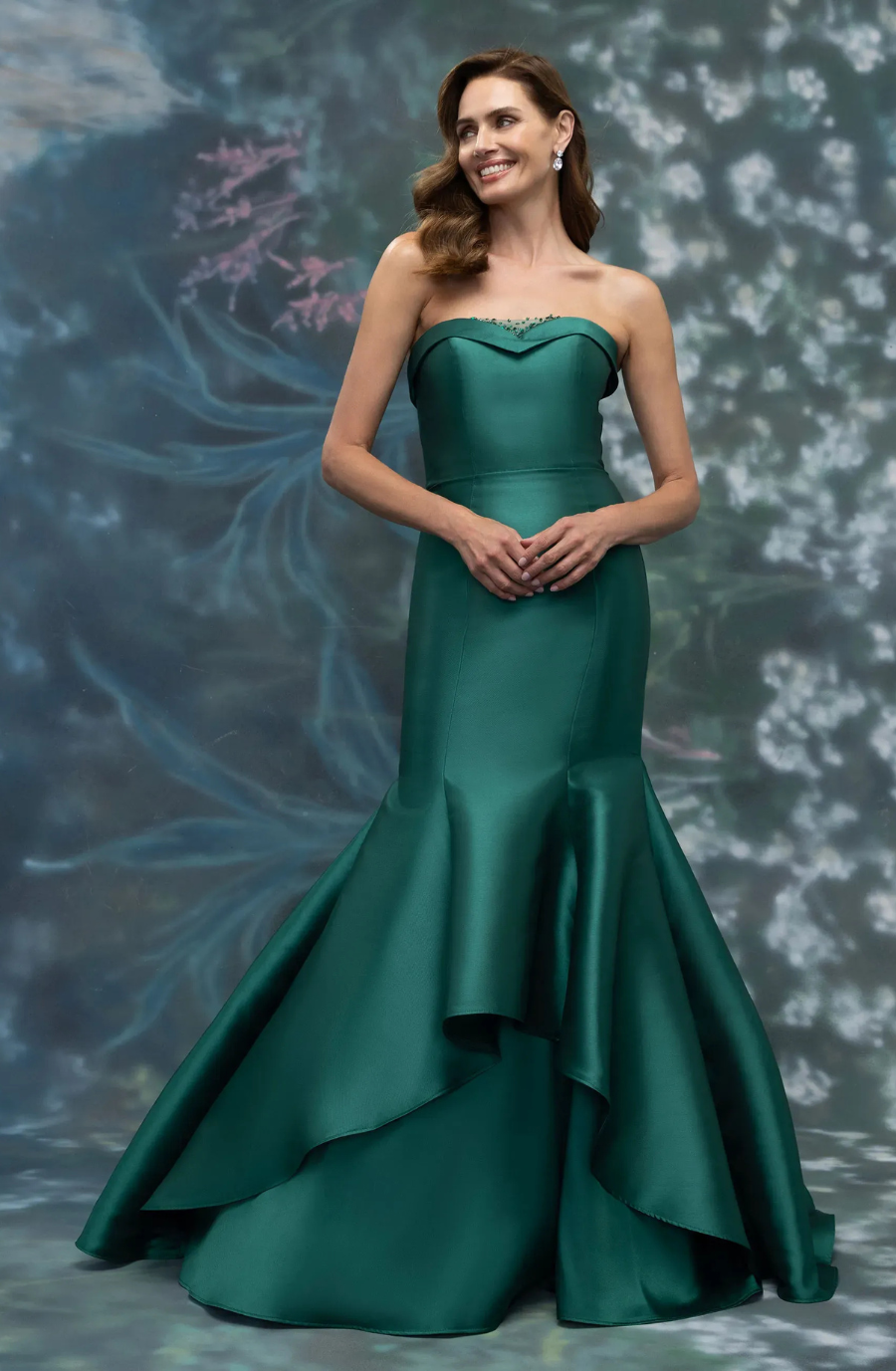 A woman wearing a strapless, emerald green gown with a fitted bodice and a flared skirt stands against a soft, floral background.